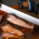 A person using a large brisket knife to slice into a partially sliced, fully cooked beef brisket flat on a cutting board.