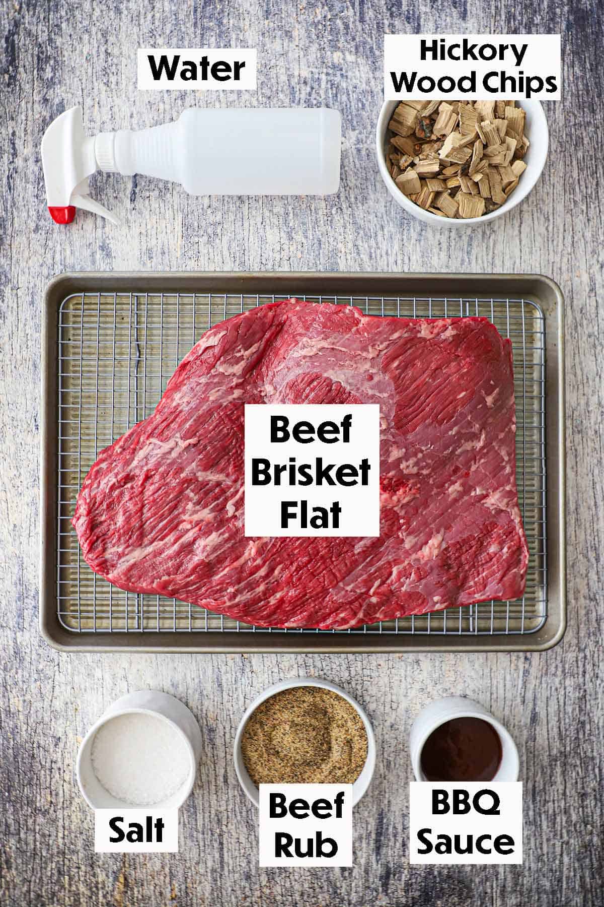 An arrangement of ingredients for Texas-Style Smoked Brisket on a grey wooden background including an uncooked brisket flat on a baking rack in a baking sheet, Hickory wood chips, salt, beef run, and barbecue sauce.