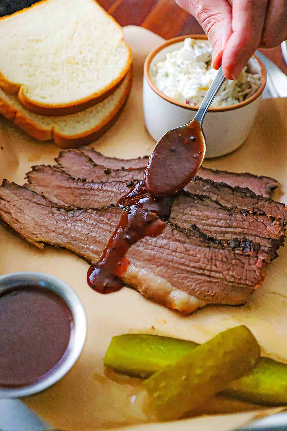 A person drizzling barbecue sauce across the center of three strips of Texas-Style Smoked Brisket that is on a trey lined with brown paper and is surrounded by white bread slices, a bowl of coleslaw, a small bowl of BBQ sauce, and two pickle spears.