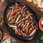 An overhead view of veggie fajitas on a large fajita skillet resting on a wooden board with charred flour tortillas resting nearby.
