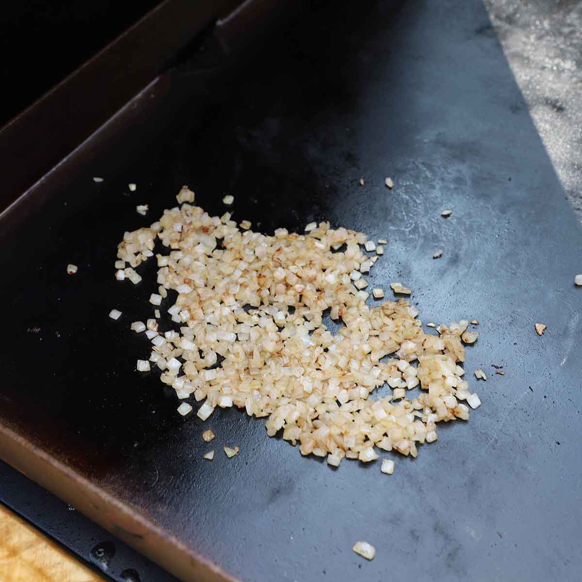 A pile of chopped yellow onions being sautéed in a little oil on a large outdoor griddle.