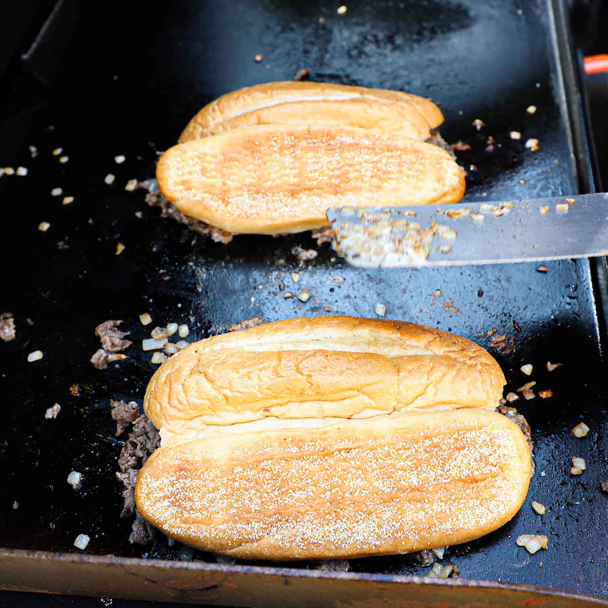 A person that has just placed two large hoagie buns over a mound of Philly cheesesteak piles on a sizzling outdoor griddle.