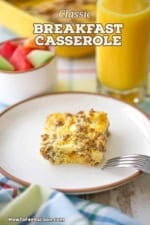 A square piece of classic breakfast casserole on a white plate next to a glass of orange juice and a small bowl of freshly cut fruit.
