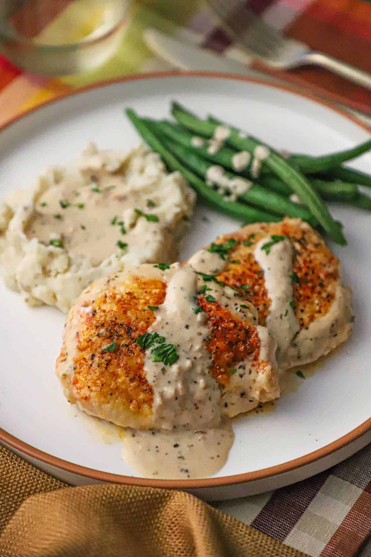 A white dinner plate filled with a serving of creamy garlic parmesan chicken with sauce drizzled over the tops of the filets resting next to a pile of mashed potatoes and steamed string beans.