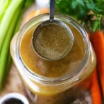An overhead view of a large ladle of homemade roasted turkey stock being held over a large jar filled with more turkey stock.