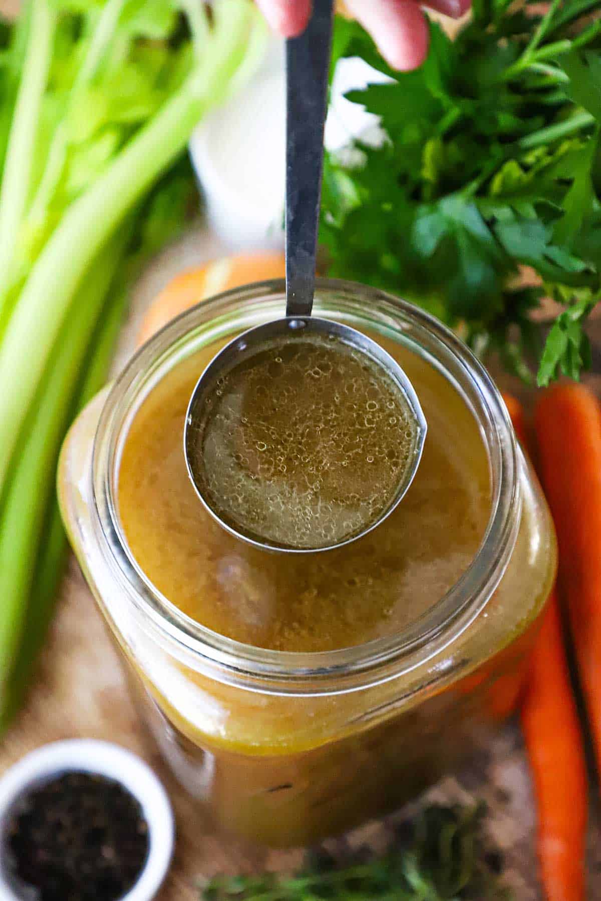 An overhead view of a large ladle of homemade roasted turkey stock being held over a large jar filled with more turkey stock.