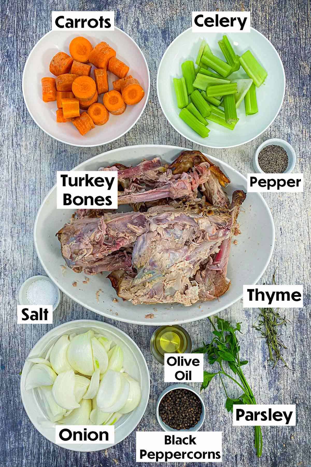 An arrangement of ingredients for homemade roasted turkey stock on a grey wooden background including turkey bone, carrots, celery, onion, olive oil, herbs, and spices.