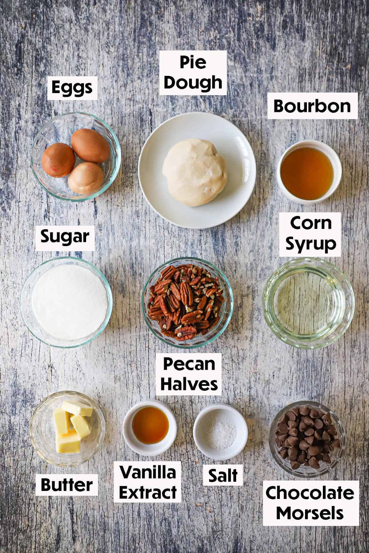 An arrangement of ingredients for the best chocolate bourbon pecan pie on a wooden background including pecans, pie dough, eggs, bourbon, chocolate, sugar, butter, corn syrup, and vanilla extract. 