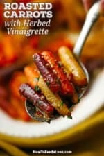 A person using a silver serving spoon to lift up a serving of roasted baby carrots with herb vinaigrette above a platter of the same.