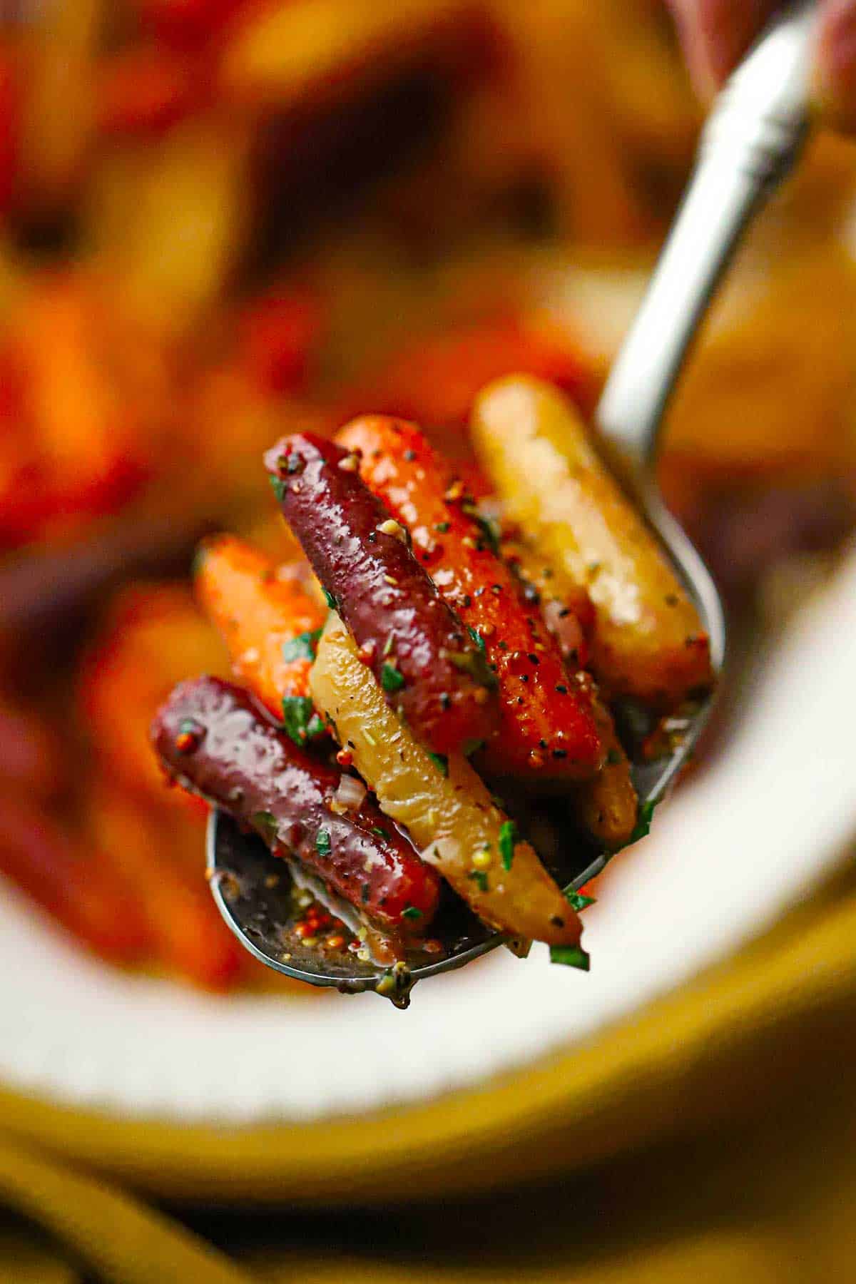 A person using a silver serving spoon to lift up a serving of roasted baby carrots with herb vinaigrette above a platter of the same.