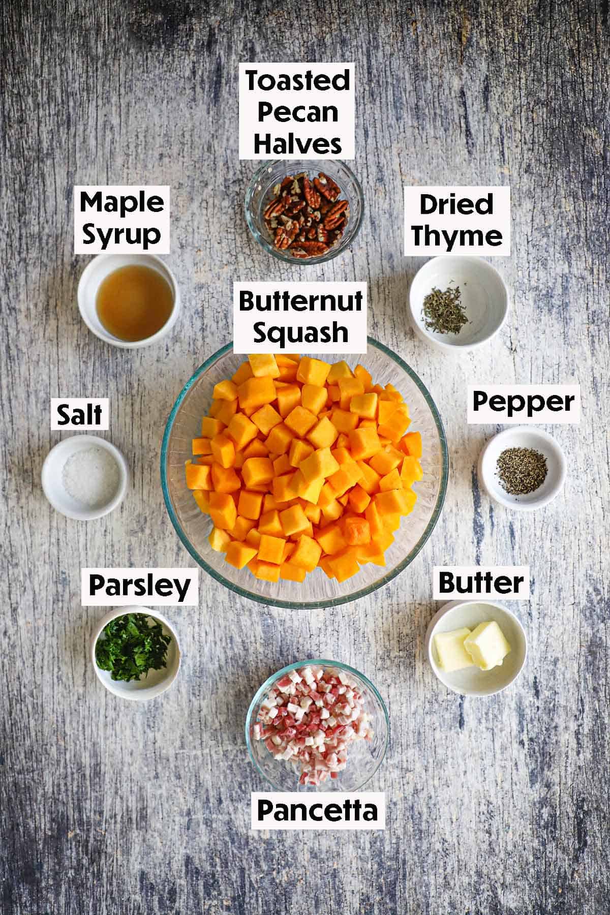 An arrangement of ingredients for maple-glazed butternut squash on a wooden grey background including cubed butternut squash, pancetta, maple syrup, butter, pecans, spices, and herbs.