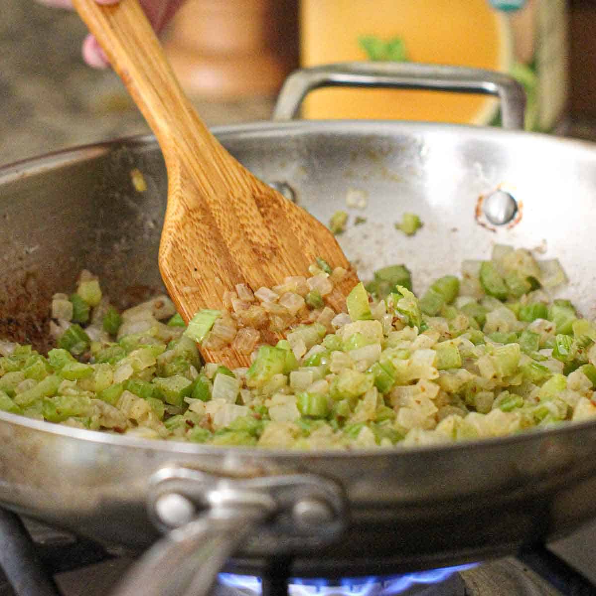 A person using a wooden spatula to stir chopped onions and celery that are being sauteed in a skillet on a gas stove.