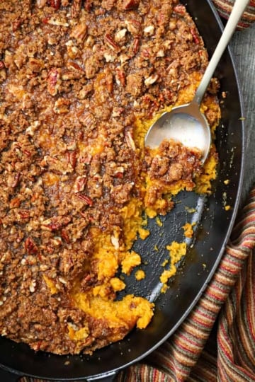 An overhead view of a sweet potato casserole with crunchy pecan topping in an oval baking dish with a portion of the casserole missing and a serving spoon inserted into the dish.