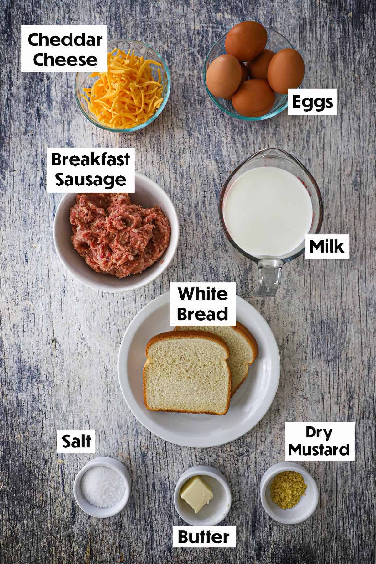 An arrangement of ingredients for classic breakfast casserole on a grey wooden background including uncooked breakfast sausage, shredded cheddar cheese, milk, two sliced of bread, dry mustard, salt, and pepper. 