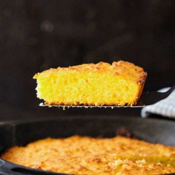 A person using a metal spatula to raised a slice of homemade cornbread from a cast-iron skillet filled with freshly baked cornbread.