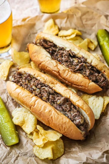 Two homemade Philly cheesesteaks sitting next to each other on wrinkled brown paper with kettle chips, pickle spears, and two beers resting near the sandwiches.