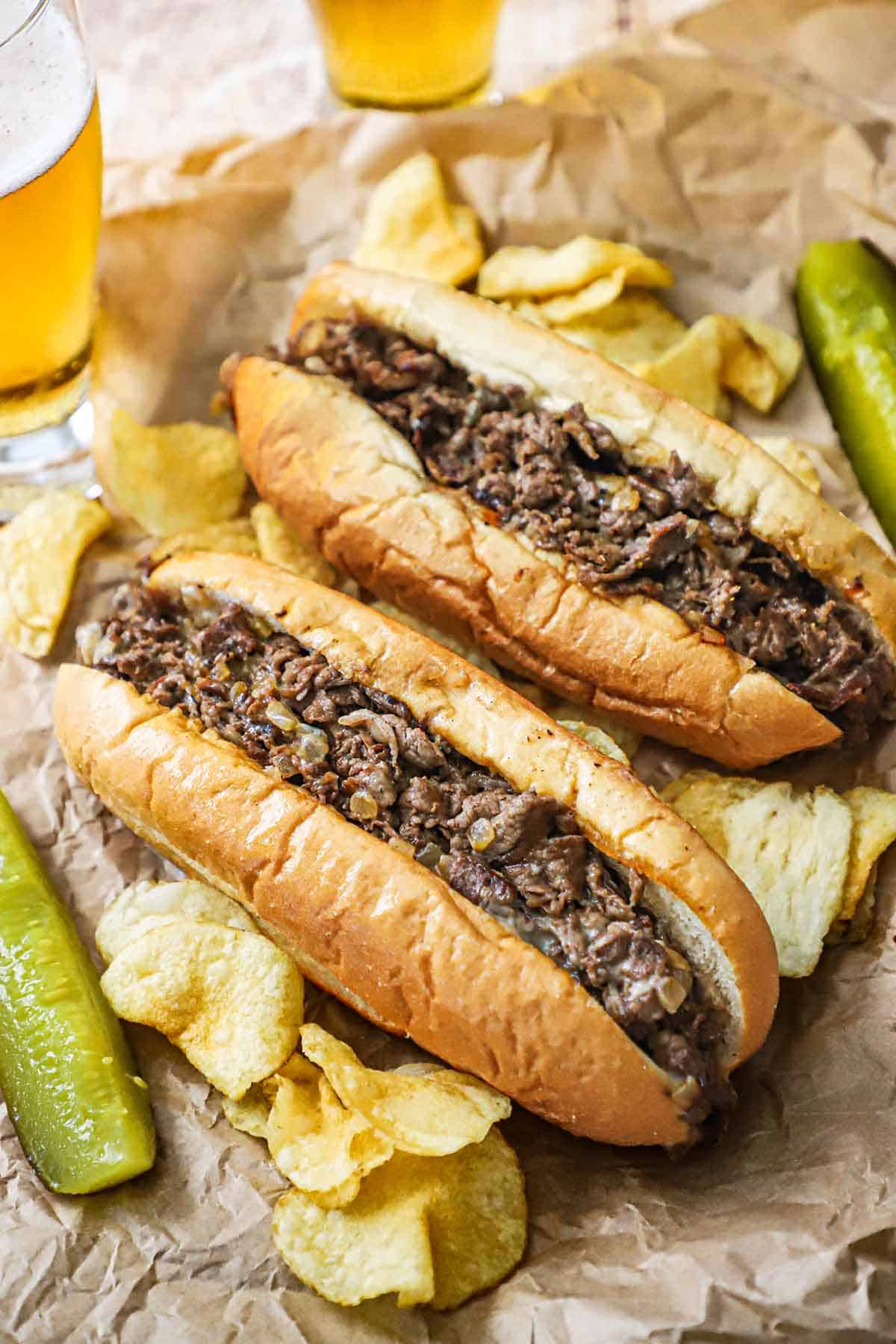 Two homemade Philly cheesesteaks sitting next to each other on wrinkled brown paper with kettle chips, pickle spears, and two beers resting near the sandwhiches.