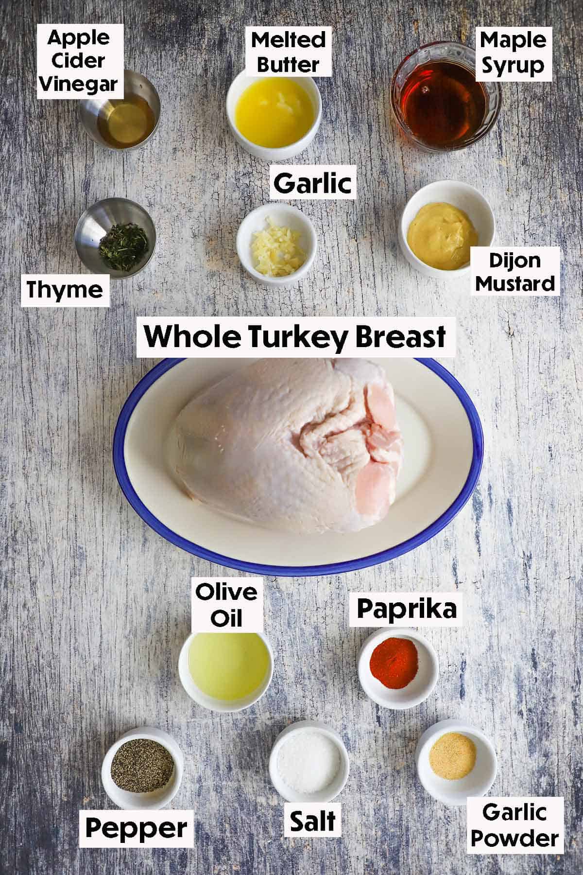 An arrangement of ingredients for a maple glazed turkey breast on a grey wooden background including an uncooked turkey breast, apple cider vinegar, butter, maple syrup, garlic, Dijon mustard, oil, herbs, and spices.