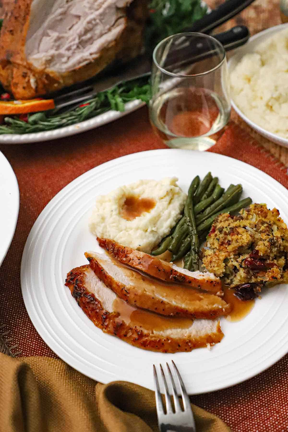 A white dinner plate filled with three slices of maple glazed turkey breast next to cornbread dressing, green bans, and mashed potatoes, all topped with turkey gravy.