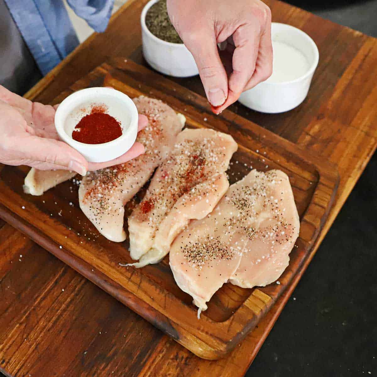 A person using his fingers to sprinkle smoked paprika over uncooked boneless chicken breasts that have already been seasoned with salt and pepper.