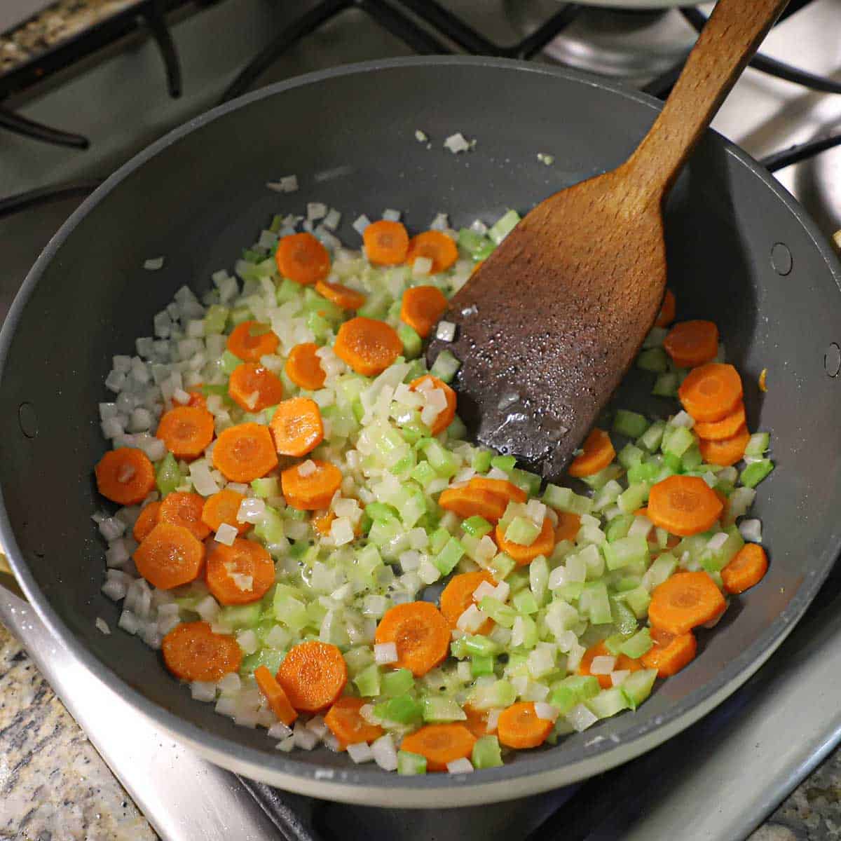 A person using a large wooden spatula to sauté chopped onions, celery, and sliced carrots in butter in a large non-stick skillet on a gas stove.