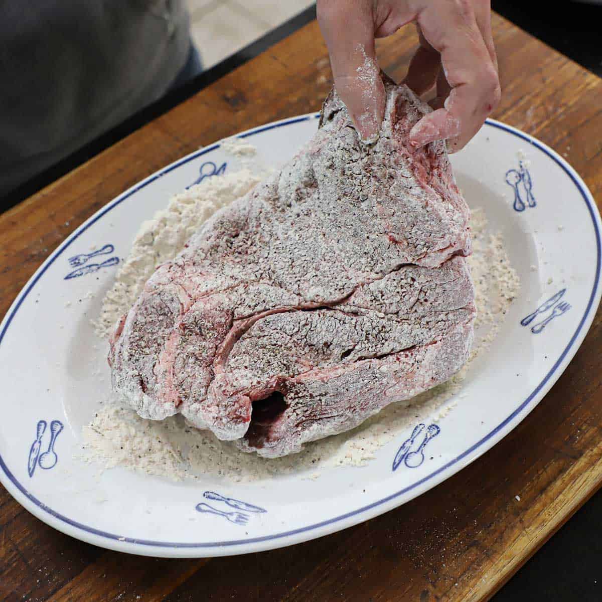 A person dredging an uncooked chuck roast through seasoned flour on a large platter.
