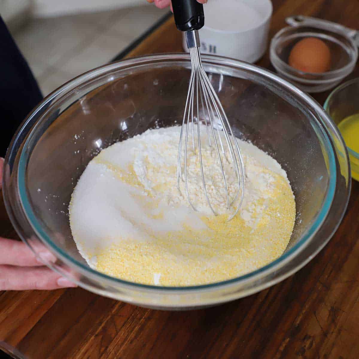 A person using a whisk to combine dry ingredients of cornmeal, flour, sugar, baking powder, and salt in a glass bowl.