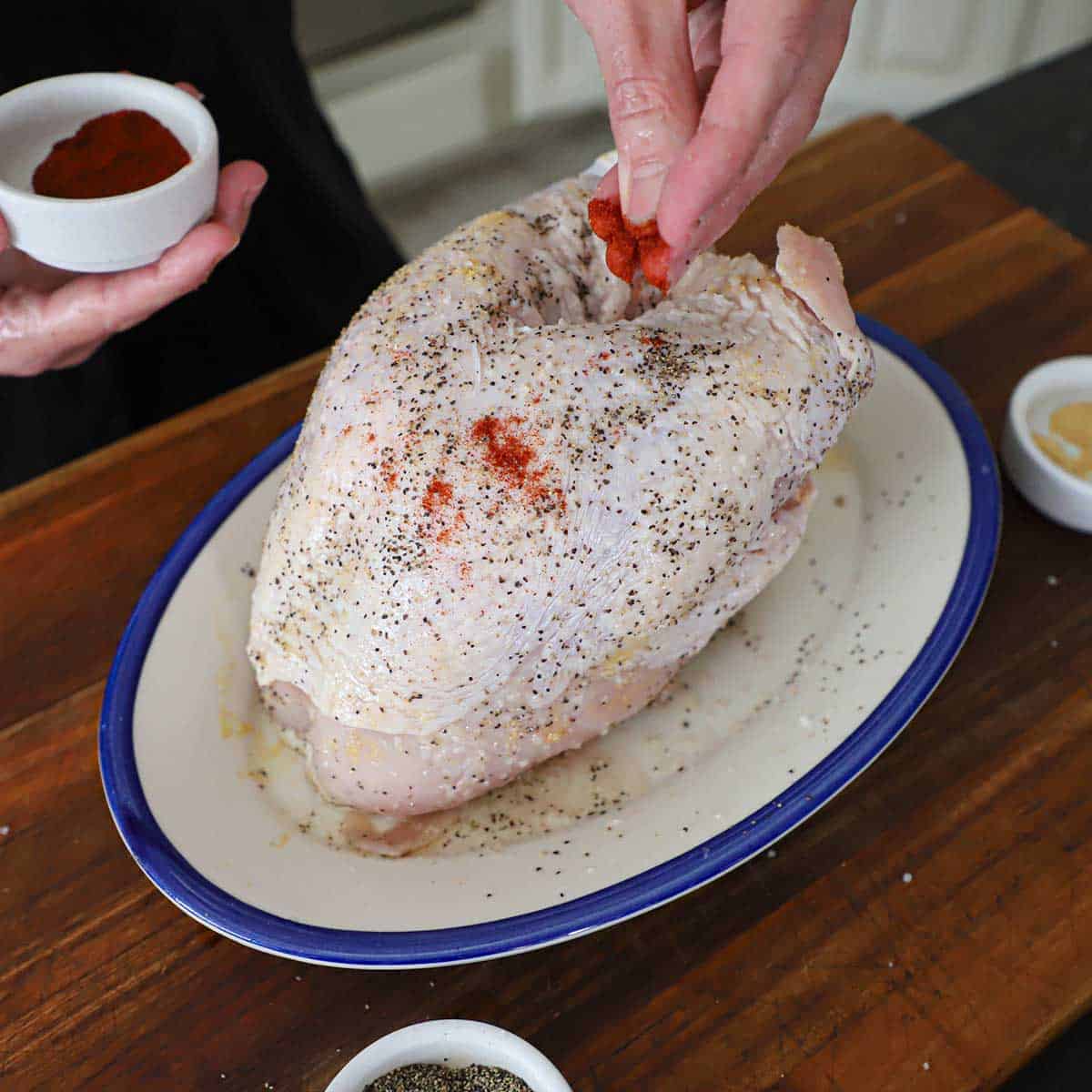 A person using his fingers to sprinkle smoked paprika over an uncooked whole turkey breast on a platter that has already been seasoned with salt and pepper.