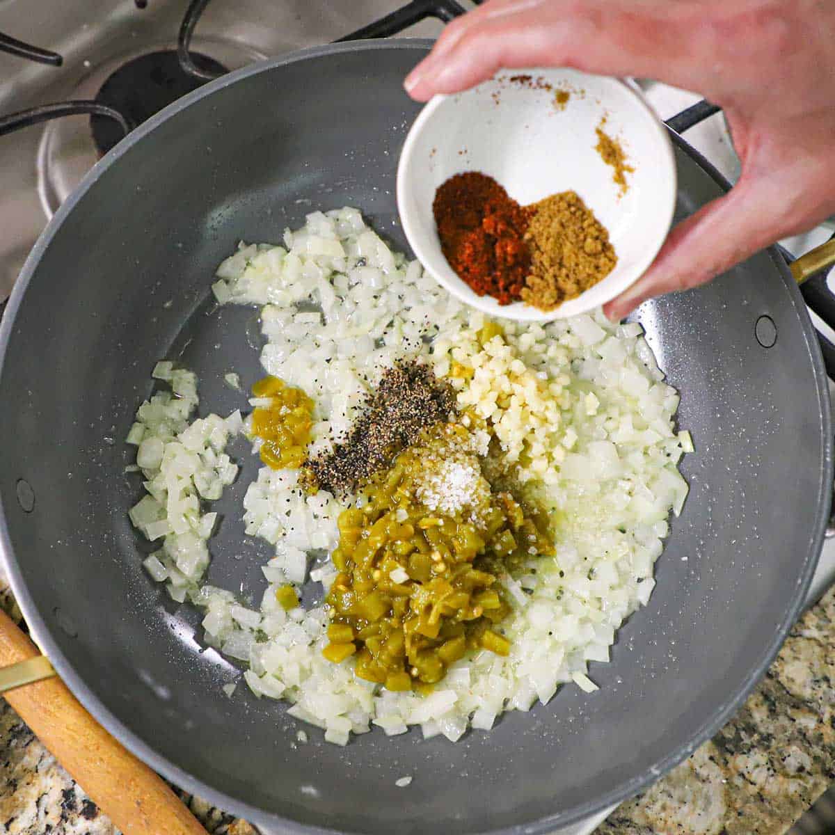 A person transferring small piles of ground chili powder and ground cumin from a small bowl into a skillet filled with sautéed onions, green chiles, chopped garlic, and other seasonings.