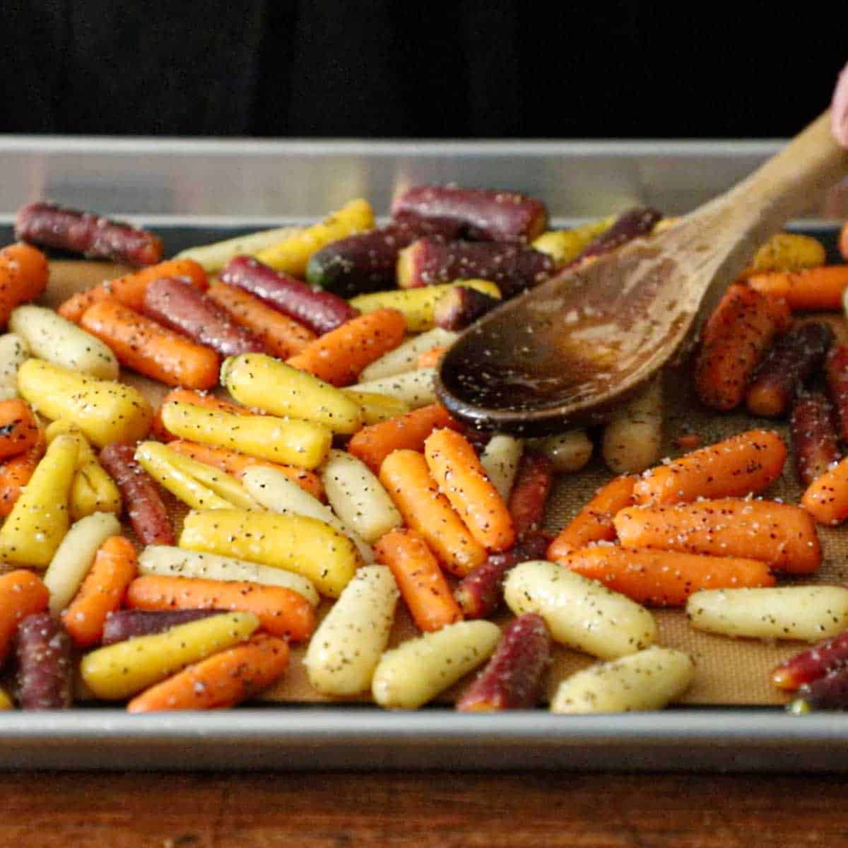 A person using a wooden spoon to spread lightly seasoned baby carrots in a single row on a metal baking sheet lined with a silicone baking matt.