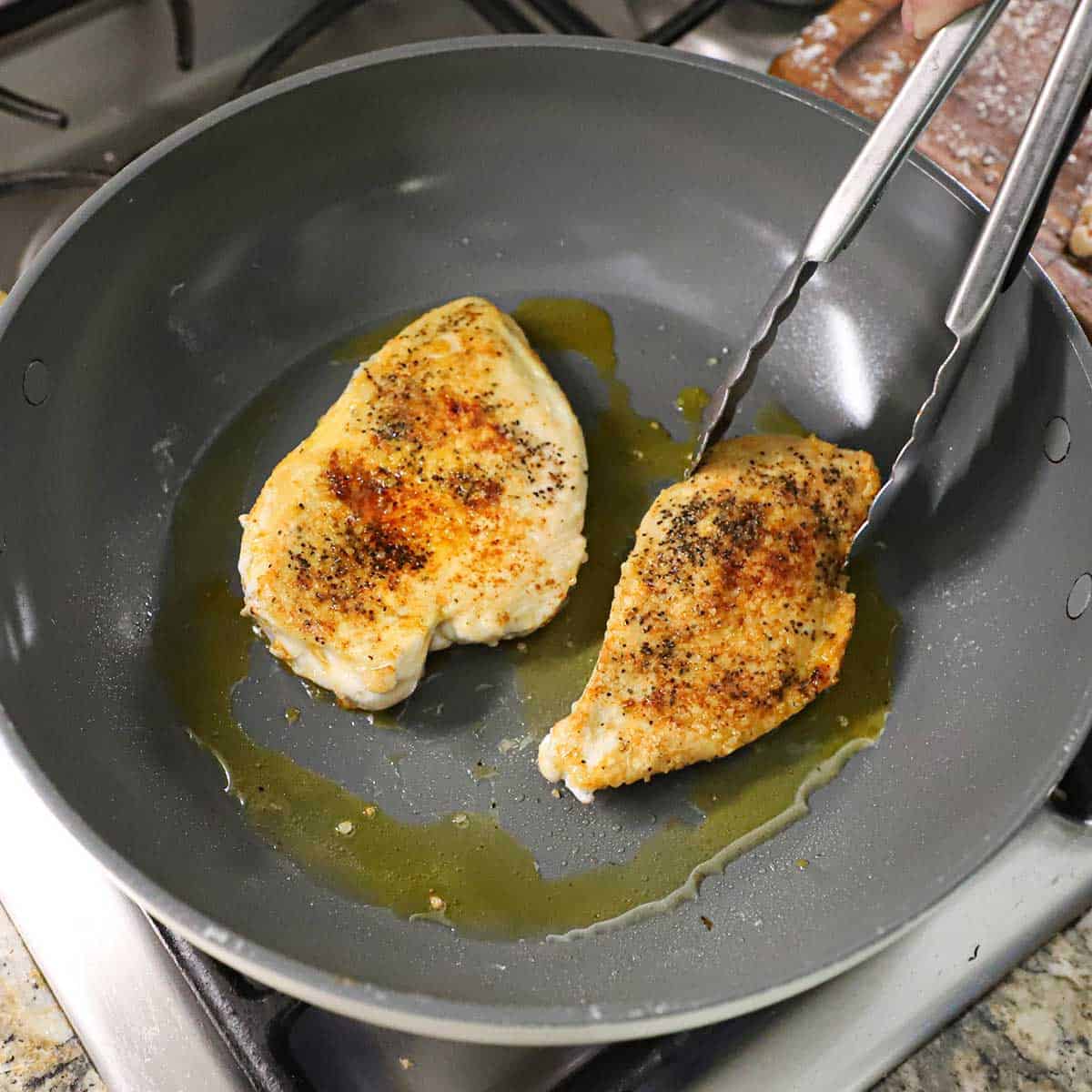 A person using a pair of metal tongs to flip a seared seasoned chicken breast in a non-stick skillet with a second seared breast simmering in a thin layer of vegetable oil.