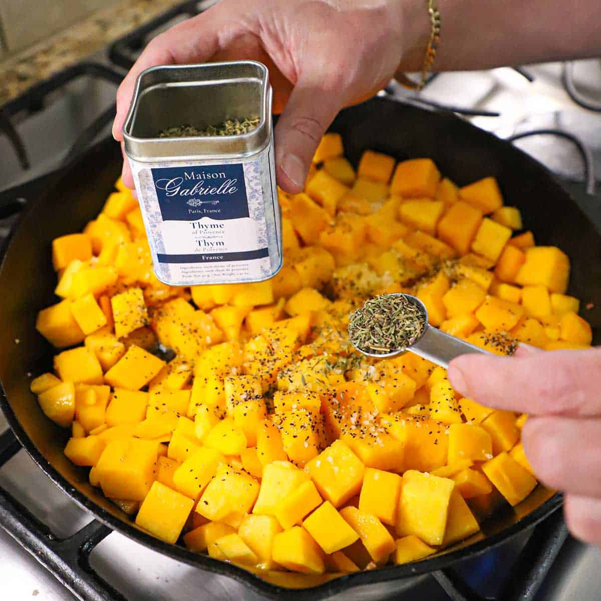 A person holding a metal tin of dried thyme with one hand over a skillet filled with cubed butternut squash and the other hand holding a teaspoon of the dried herb.