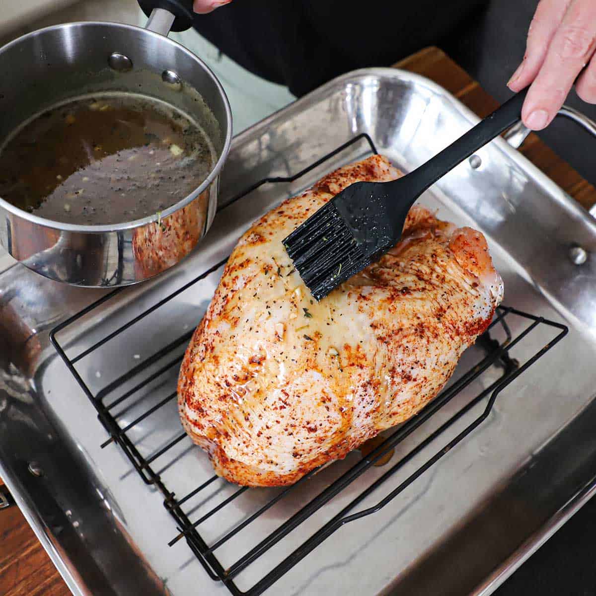 A person using a small black brush to apply a maple glaze to a whole turkey breast that is resting on a baking rack in a roasting pan that is about to go into the oven.