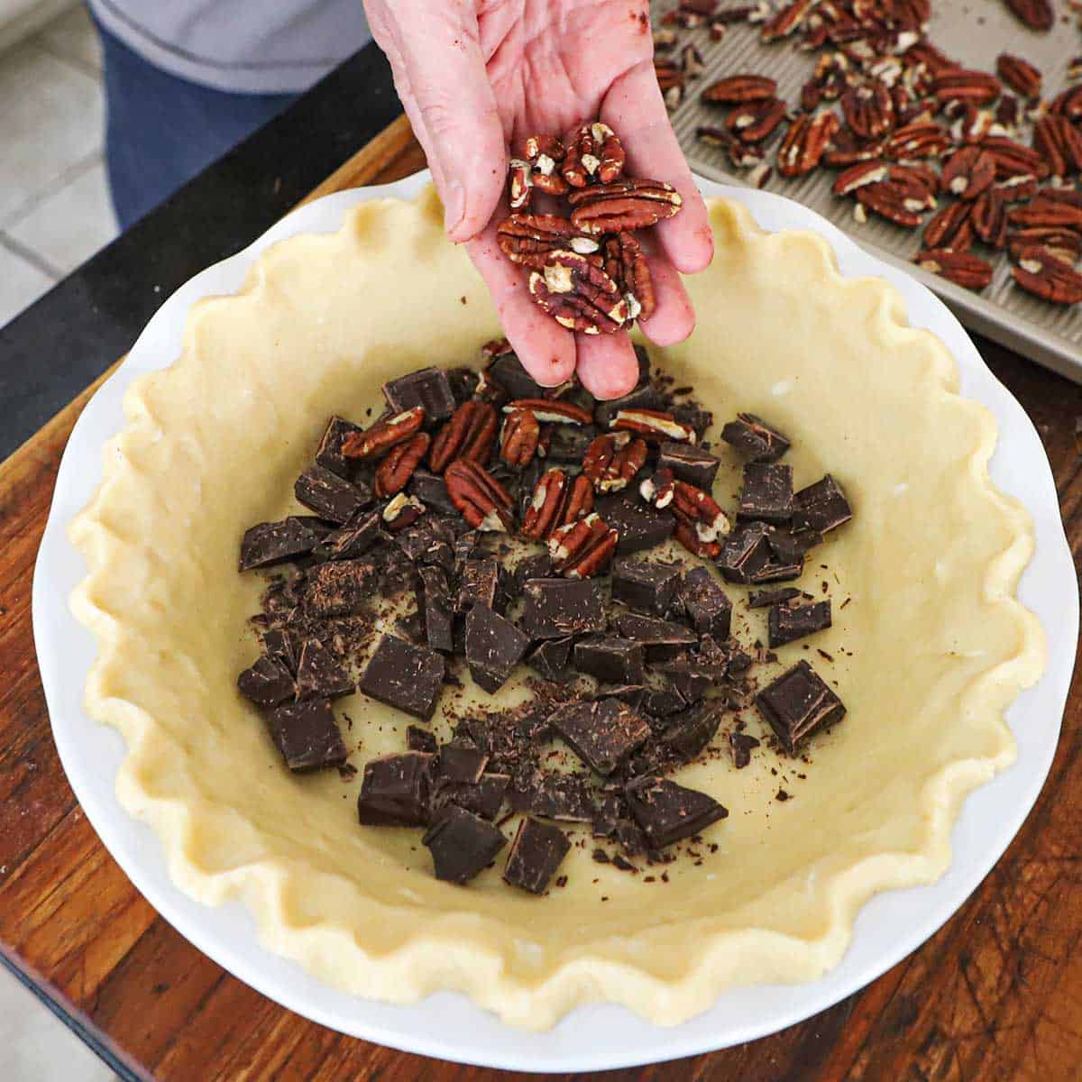 A person sprinkling toasted pecan halves over chunks of chocolate in the bottom of an uncooked pie dough that has been fitted into a white pie dish.