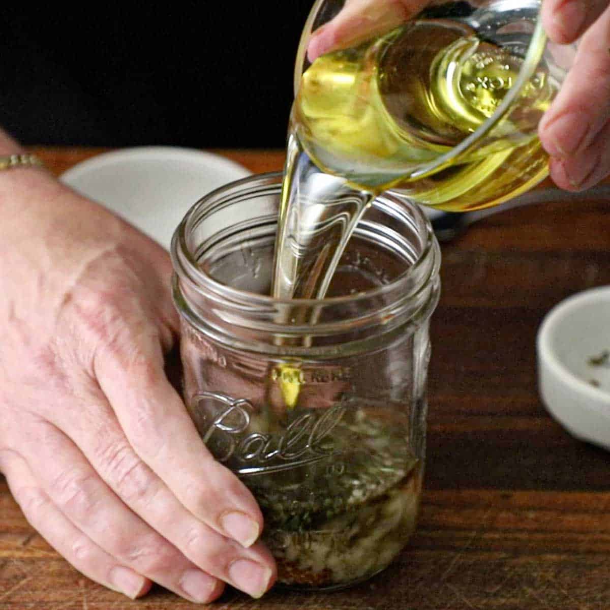 A person pouring olive oil from a small glass bowl into a jar filled with white wine vinegar, whole grain mustard, minced garlic, and herbs.