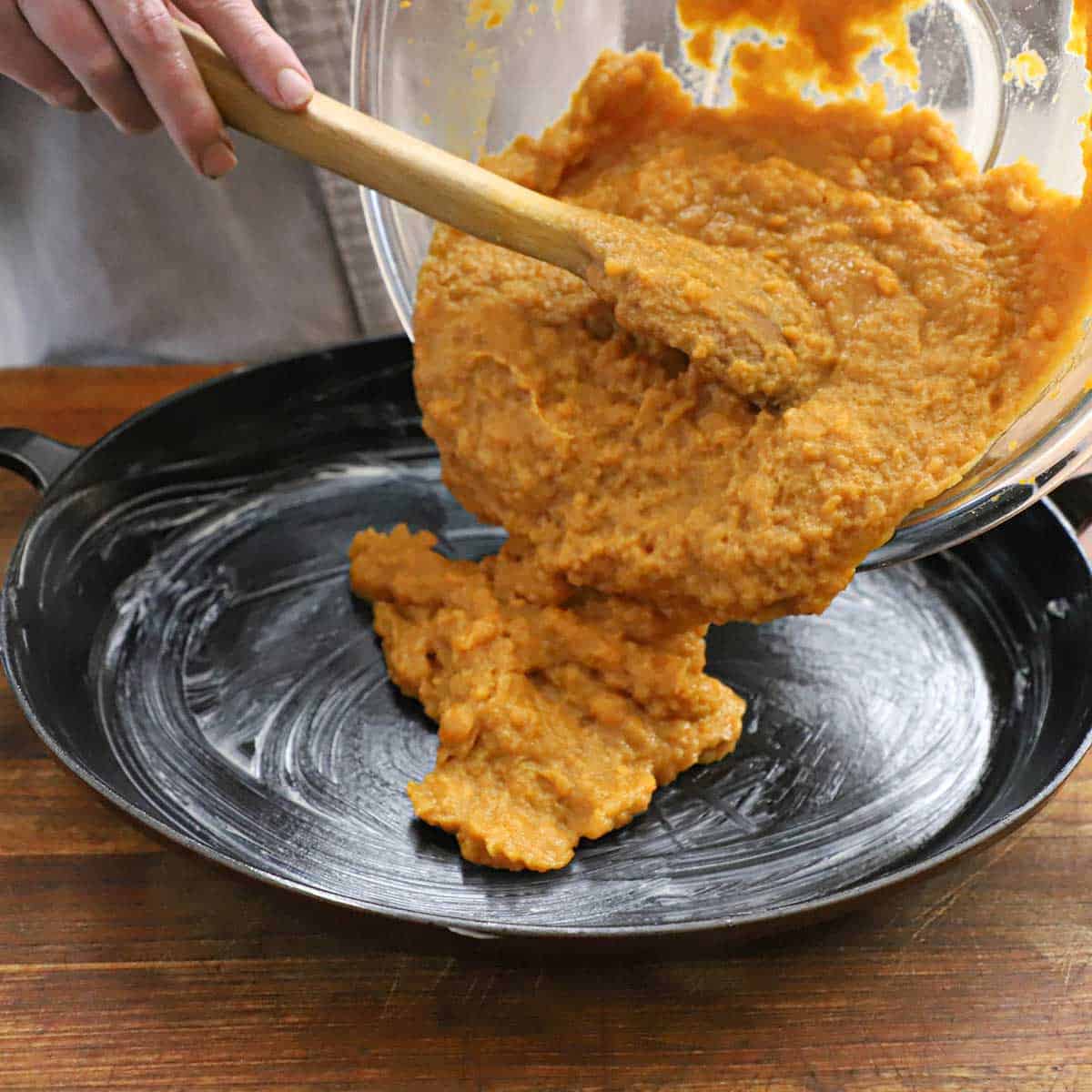 A person transferring a sweet potato casserole mixture with a wooden spoon into a greased baking dish.