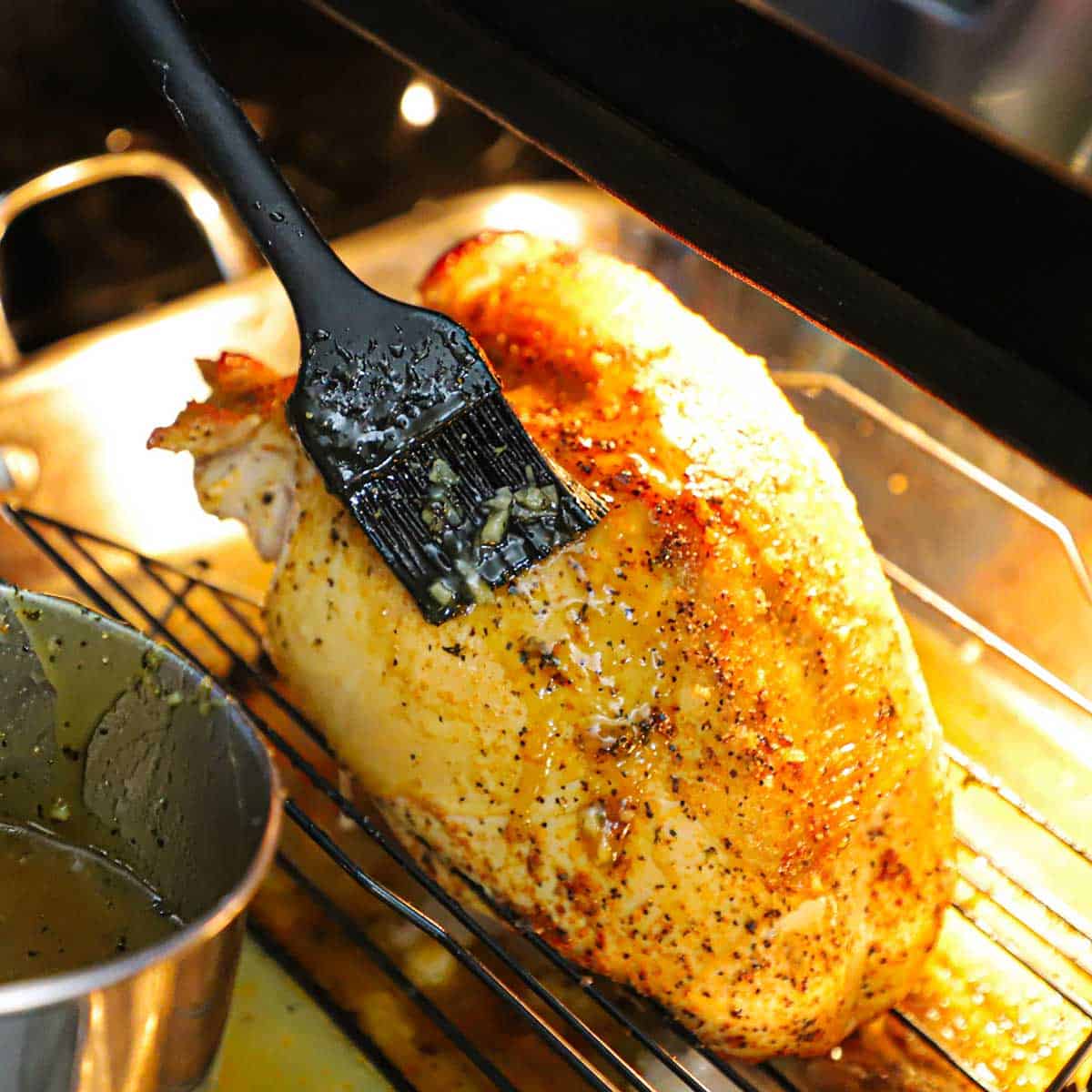 A person using a small black brush to baste a partially roasted whole turkey breast with a maple glaze on a baking rack in a roasting pan inside an oven.