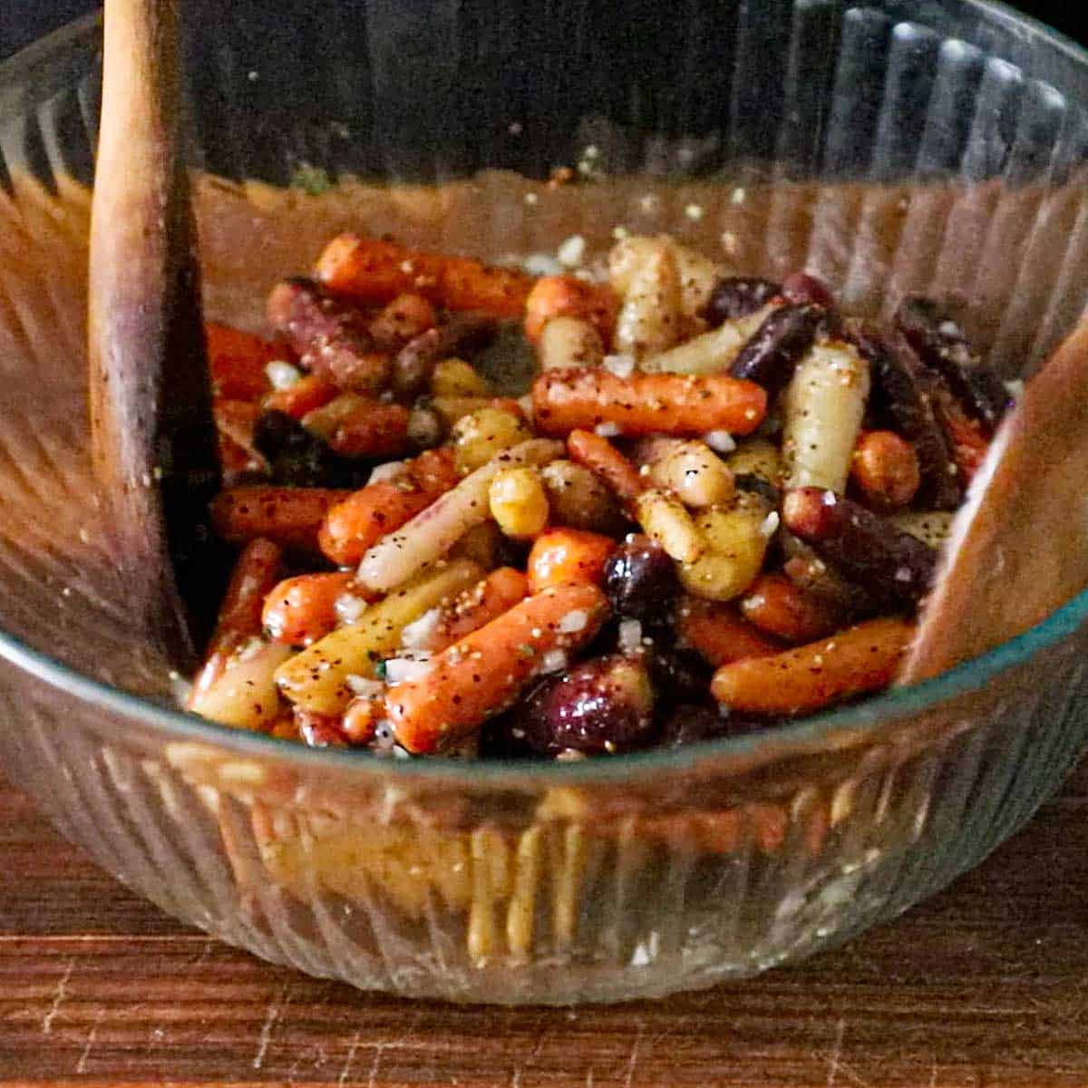 A person using two wooden spatula to toss together roasted baby carrots that are coated with an herbed vinaigrette all in a large glass bowl.