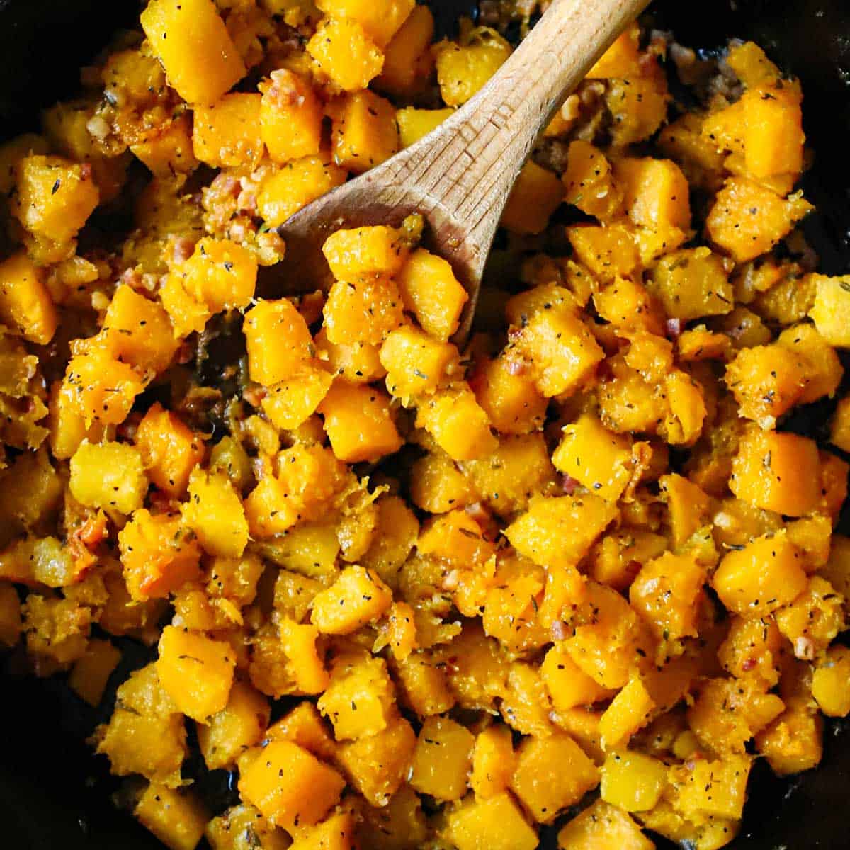 A wooden spoon inserted into the center of a skillet filled with Maple-Glazed Butternut Squash with Pancetta and Herbs.
