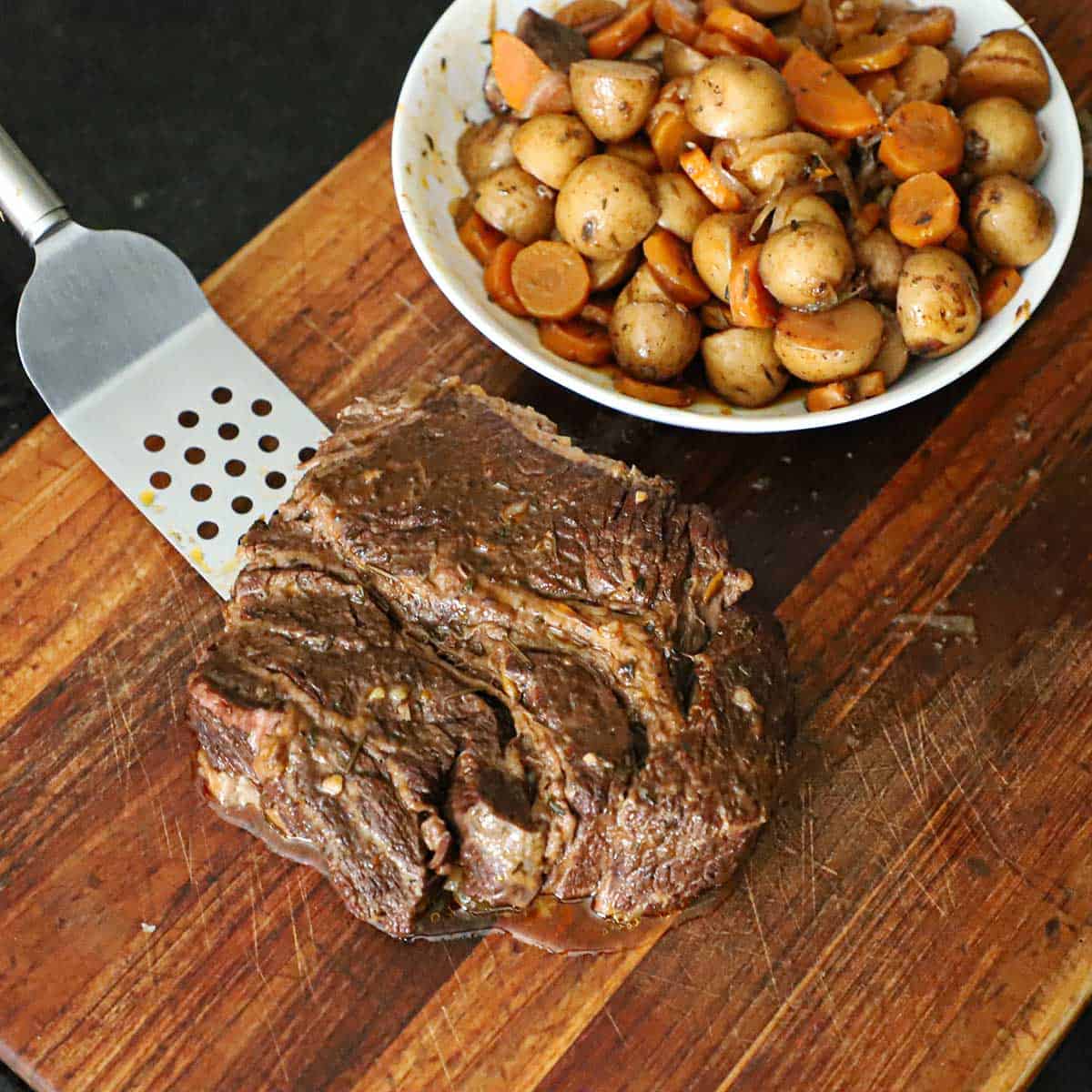 A person using a metal spatula to place a slow cooker pot roast onto a wooden cutting board next to a bowl of slow cooked vegetables.