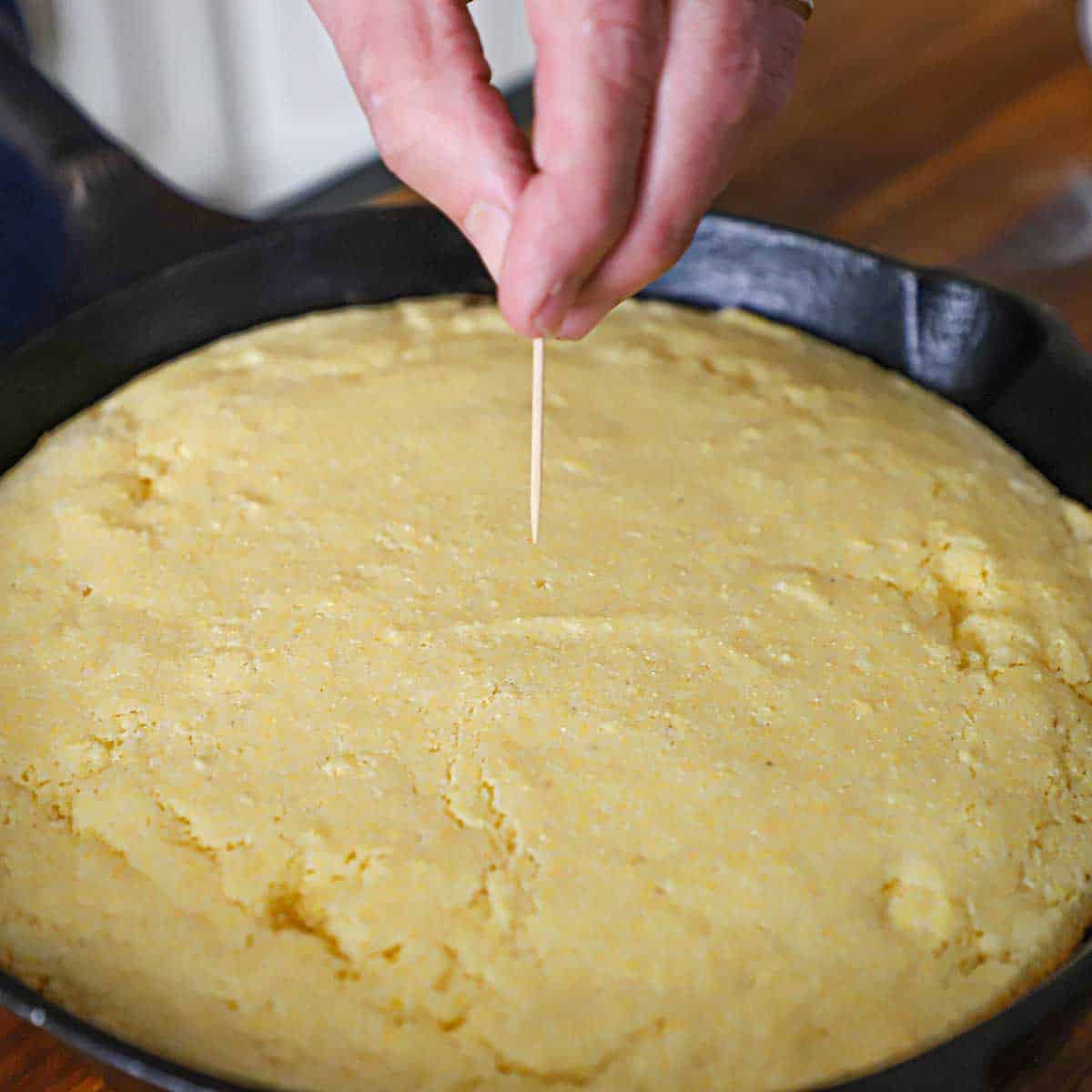 A person pulling a dry toothpick from a freshly baked pan of homemade cornbread in a large cast-iron skillet.