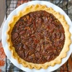 An overhead view of the best chocolate bourbon pecan pie in a white pie dish resting on a baking rack on an autumnal linen underneath.