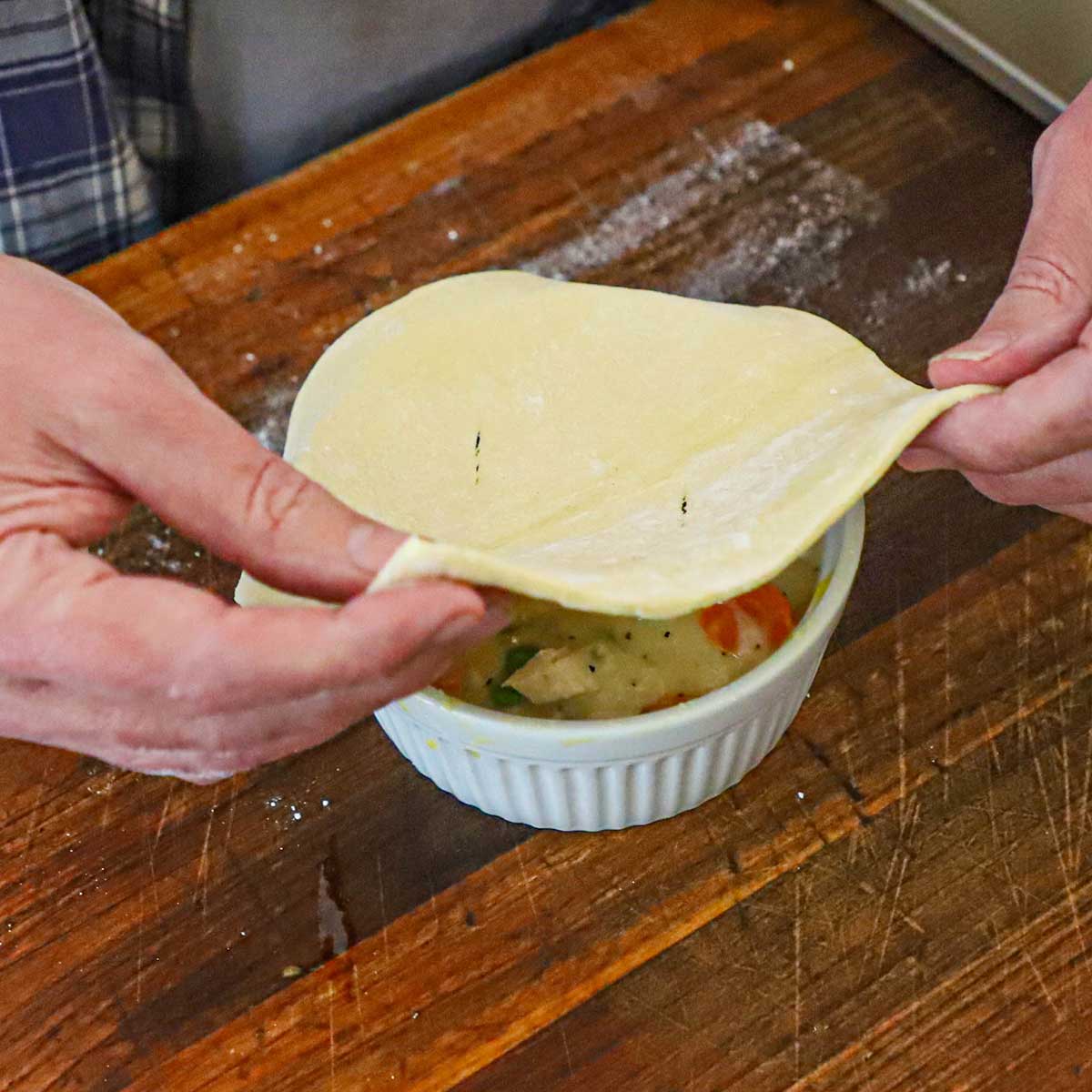 A person placing a square puff pastry over the top of a ramekin that is filled with leftover turkey pot pie filling.