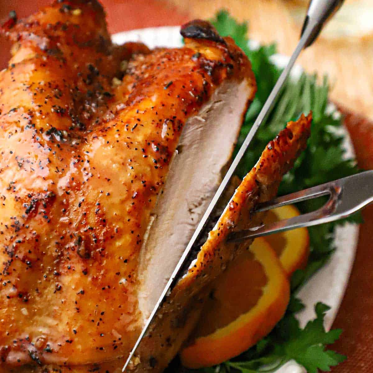 A person using a carving fork and knife to slice into a freshly roasted maple glazed turkey breast that is resting on a platter surrounded by fresh herbs and sliced oranges.