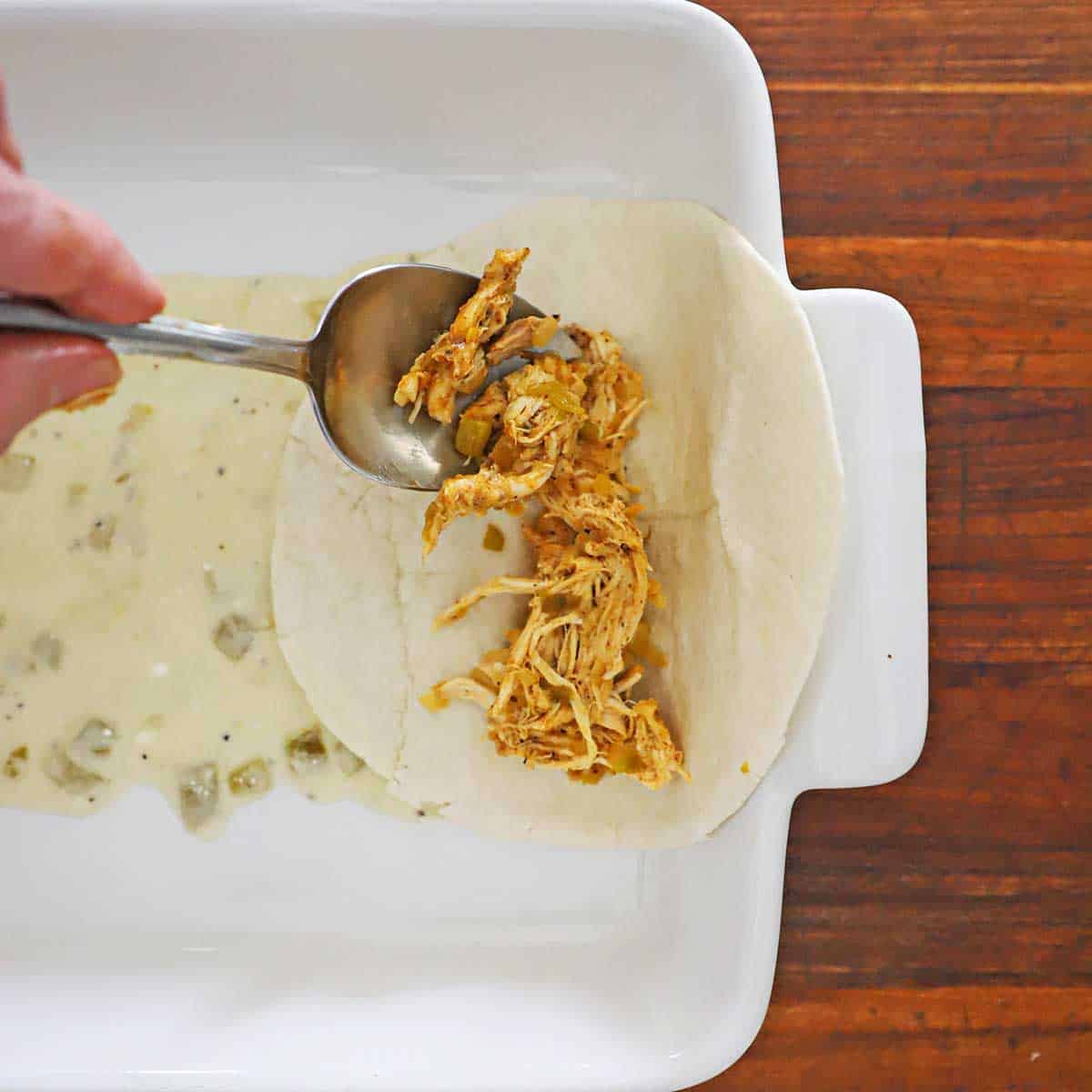 A person using a spoon to transfer chicken enchilada meat into the center of a flour tortilla the is placed in casserole dish with a thin layer of a sour cream sauce in the bottom of the dish.