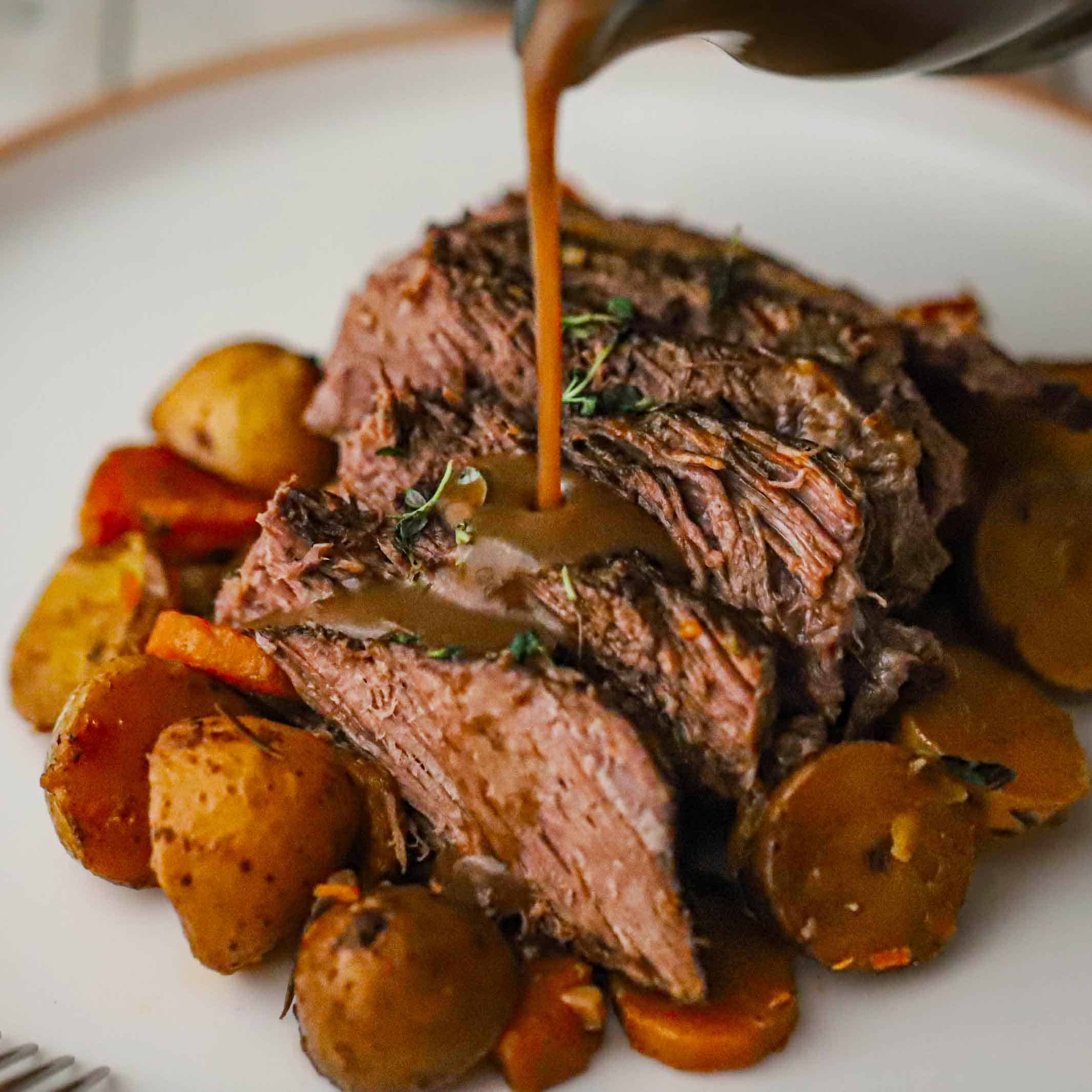 Brown gravy being poured over slices of the best slow cooker pot roast on a white dinner plate also holding cooked potatoes and carrots.