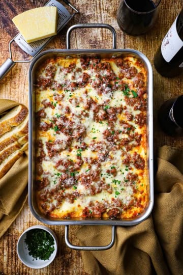 An overhead view of a steel lasagna pan filled from end to end with the best weeknight lasagna with a block parmesan cheese and a bottle of red wine nearby.