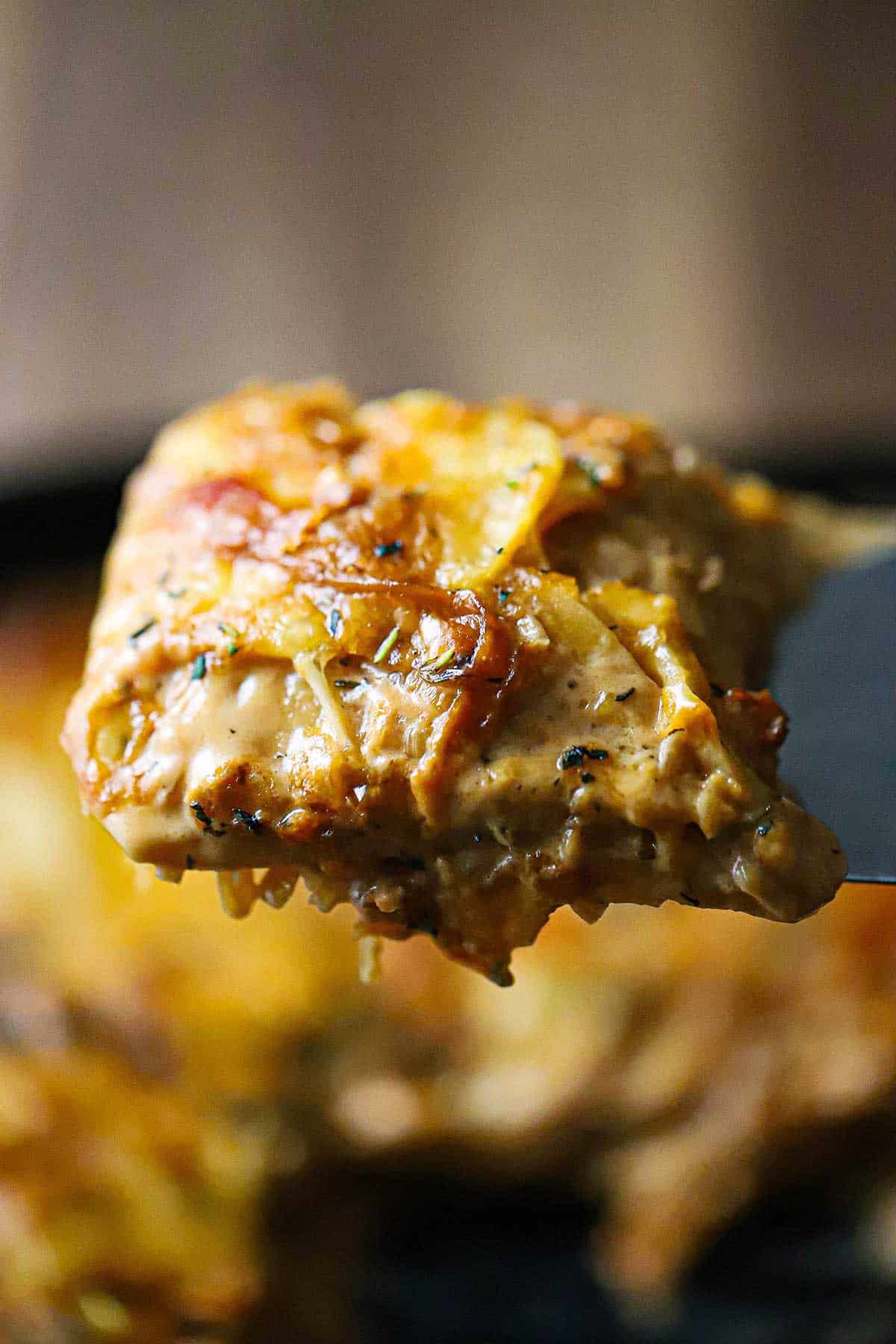 A person using a large metal spatula to raise up a square piece of French onion au gratin potatoes over a skillet filled with the same.