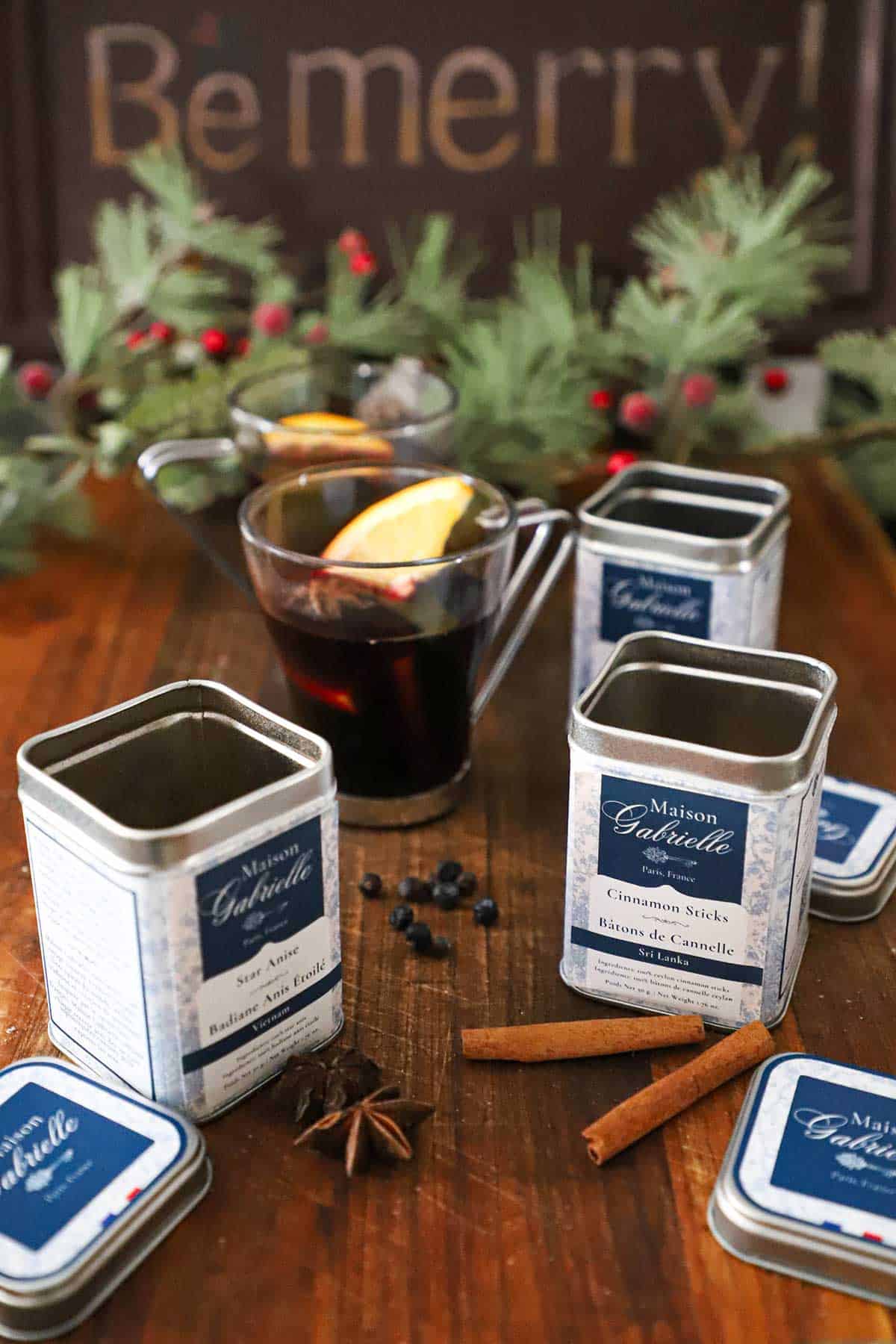 Several tins of spices sitting around a glass mug filled with glehwien (mulled wine) with a festive garland and sign that reads "Be Merry" behind it.