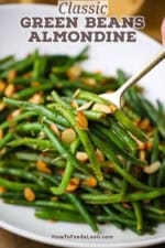 A person using a gold serving utensil to lift a serving of green beans almondine from a white platter of the dish.